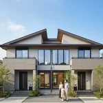 青空の下、現代的なデザインの積水ハウスの二世帯住宅の前に立つ夫婦と一人の子供の家族。家の玄関が二つあり、左右対称の構造が特徴的。